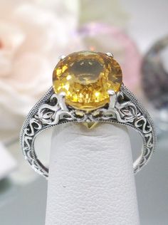 Natural Yellow Citrine Sterling Silver Ring Swan Design #D190 Introducing our Natural Yellow Citrine Swan Ring, D190, an exquisite piece that effortlessly blends elegance with vintage charm. Crafted with meticulous attention to detail, this Art Deco-inspired ring features a stunning natural yellow citrine gemstone nestled within a sterling silver filigree setting. At the heart of this ring is a full cut oval citrine, measuring 12mm in length and 10mm in width, radiating warmth and vibrancy. The intricate filigree setting adds a touch of sophistication, with delicate etched designs that evoke the timeless beauty of the Victorian era. One cannot help but be captivated by the graceful swan-like pattern adorning the sides of the band, each tiny swirl enhancing the ring's overall allure. The ri Orange Round Yellow Sapphire Jewelry, Formal Yellow Solitaire Jewelry, Yellow Sapphire Topaz Ring For Wedding, Yellow Topaz Ring With Center Stone For Formal Occasions, Elegant Yellow Ring With Center Stone, Formal Yellow Topaz Ring With Center Stone, Formal Oval Yellow Topaz Ring, Formal Yellow Oval Topaz Ring, Yellow Round Topaz Ring With Prong Setting