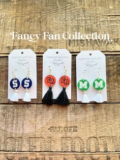 three pairs of earrings with tassels and letters on them, hanging from tags