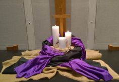 three white candles sitting on top of a table next to a purple cloth and cross