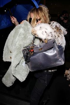 a blonde woman carrying a gray purse with white fur on it and holding an umbrella