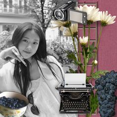 a woman sitting in front of a typewriter next to flowers and a bowl of blueberries