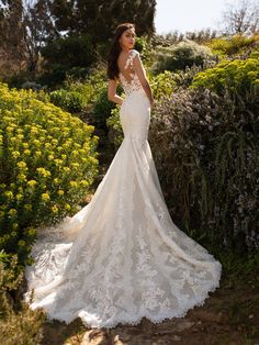 a woman in a white wedding dress standing by some bushes and flowers with her back turned to the camera