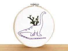 a small embroidery project with a dinosaur and plants on it's back side, sitting on a wooden surface