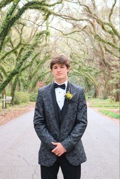 a young man in a tuxedo poses for a photo on a road lined with trees