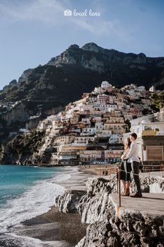 two people are standing on the edge of a cliff by the ocean with houses in the background