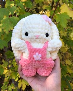 a hand holding a small crocheted white and pink stuffed animal in front of some leaves