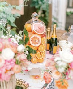 an arrangement of oranges under a clochet on a table with flowers and bottles