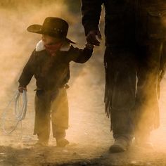 a little boy holding the hand of an adult in a cowboy hat and black outfit