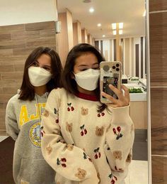 two women wearing face masks taking a selfie in front of a mirror with their cell phone