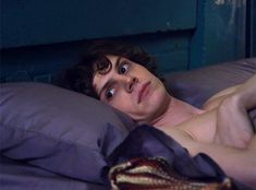 a shirtless man laying in bed with his hand on the pillow and looking at the camera