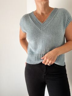 a woman standing in front of a white wall wearing a blue sweater and black pants