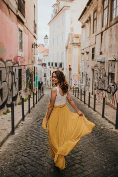 a woman is walking down the street in a yellow skirt and white tank top with her hands in her pockets