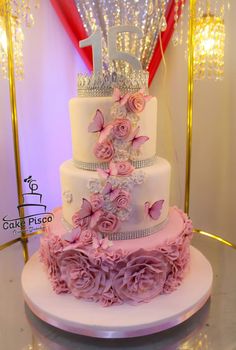 a three tiered wedding cake with pink flowers and butterflies on the top, sitting on a table in front of a chandelier