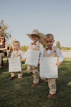 two young boys holding up signs in front of their parents on the grass at an outdoor wedding