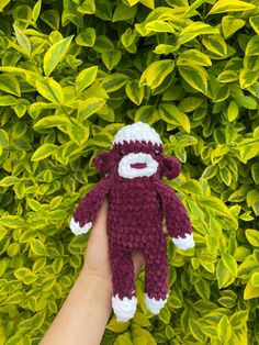 a hand holding a small stuffed animal in front of some bushes and green leaves,