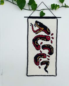 an embroidered wall hanging with a snake on it's side and green leaves in the background