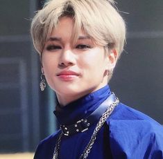 a woman with blonde hair wearing a blue shirt and silver chains on her neck is looking at the camera