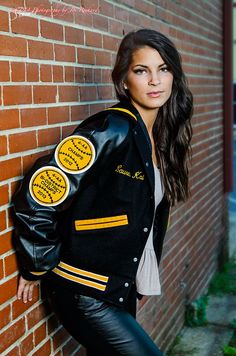 a woman leaning against a brick wall wearing a black jacket with yellow patches on it