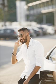 a man sitting on the hood of a car talking on his cell phone and smiling