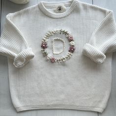 a white sweater with the letter o on it and flowers in the middle, sitting next to a ball of yarn