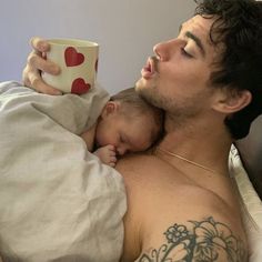 a man laying in bed holding a baby and drinking from a cup