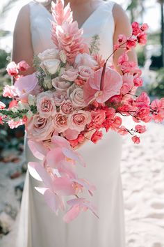 a woman in a white dress holding a pink bouquet on the beach with palm trees behind her