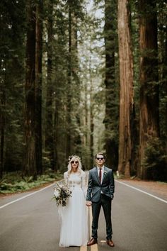 a bride and groom standing in the middle of an empty road surrounded by tall trees