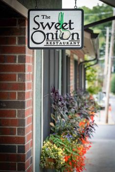 the sweet onion restaurant sign hanging from the side of a building with flowers in front of it