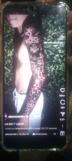 a person holding up a cell phone with tattoos on their arm and chest, in front of some bushes