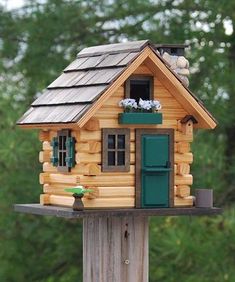 a small wooden house with green shutters and windows on top of a post in front of trees