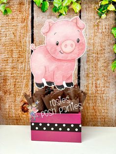 a pink pig sitting on top of a box filled with chocolates next to a wooden fence