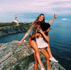 two young women sitting on top of a rock next to the ocean with their arms around each other