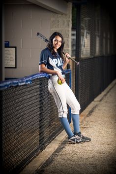 a woman holding a baseball bat and ball in her hand while leaning on a fence