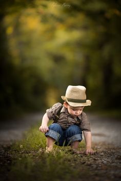 a little boy sitting in the middle of a road wearing a hat and looking down