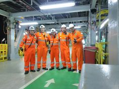 five men in orange work suits and safety vests stand together on a green floor