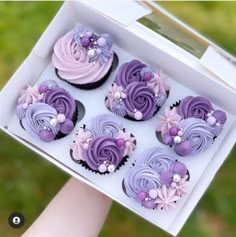 a person is holding up a box of cupcakes with purple frosting on them