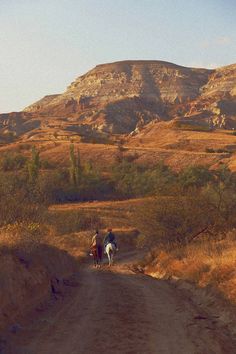 two people riding horses down a dirt road in front of a mountain with dry grass