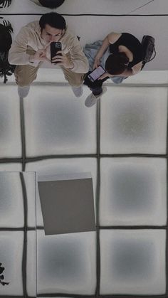 two people sitting on top of a white tiled wall with cell phones in their hands