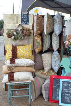 pillows and cushions are on display under an umbrella at the flea market in front of a chalkboard sign