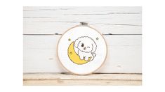 a small white dog sitting on top of a crescent moon embroidery pattern in a hoop