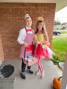 two people in costumes standing next to each other near a brick wall and pumpkins