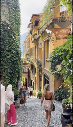 a woman walking down a cobblestone street in an old european town with lots of greenery