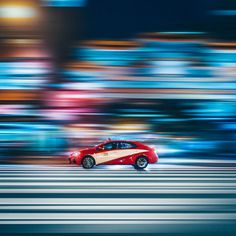 a red car is driving down the street in front of blurry buildings and trees