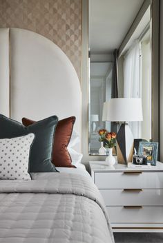 a white bed topped with lots of pillows next to a night stand and dresser under a large mirror