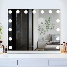 a vanity mirror with lights on it in front of a white dresser and potted plant