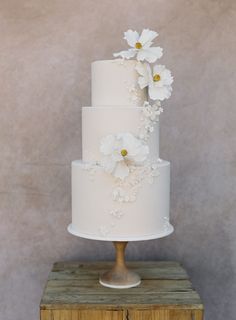 a three tiered white wedding cake with flowers on the top and bottom, sitting on a wooden stand