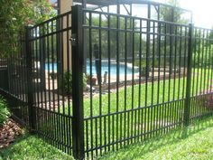 a pool behind a black fence in the grass next to some bushes and flowers near a house