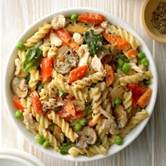 a white bowl filled with pasta and vegetables on top of a wooden table next to a spoon