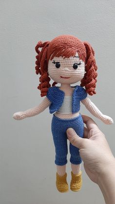 a hand holding a small doll made out of knitted material with red hair and blue pants