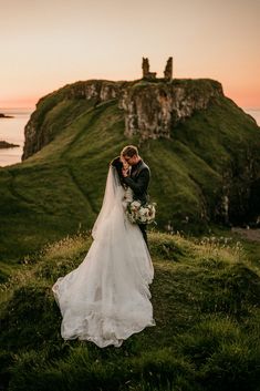 a bride and groom standing on top of a grass covered hill next to the ocean